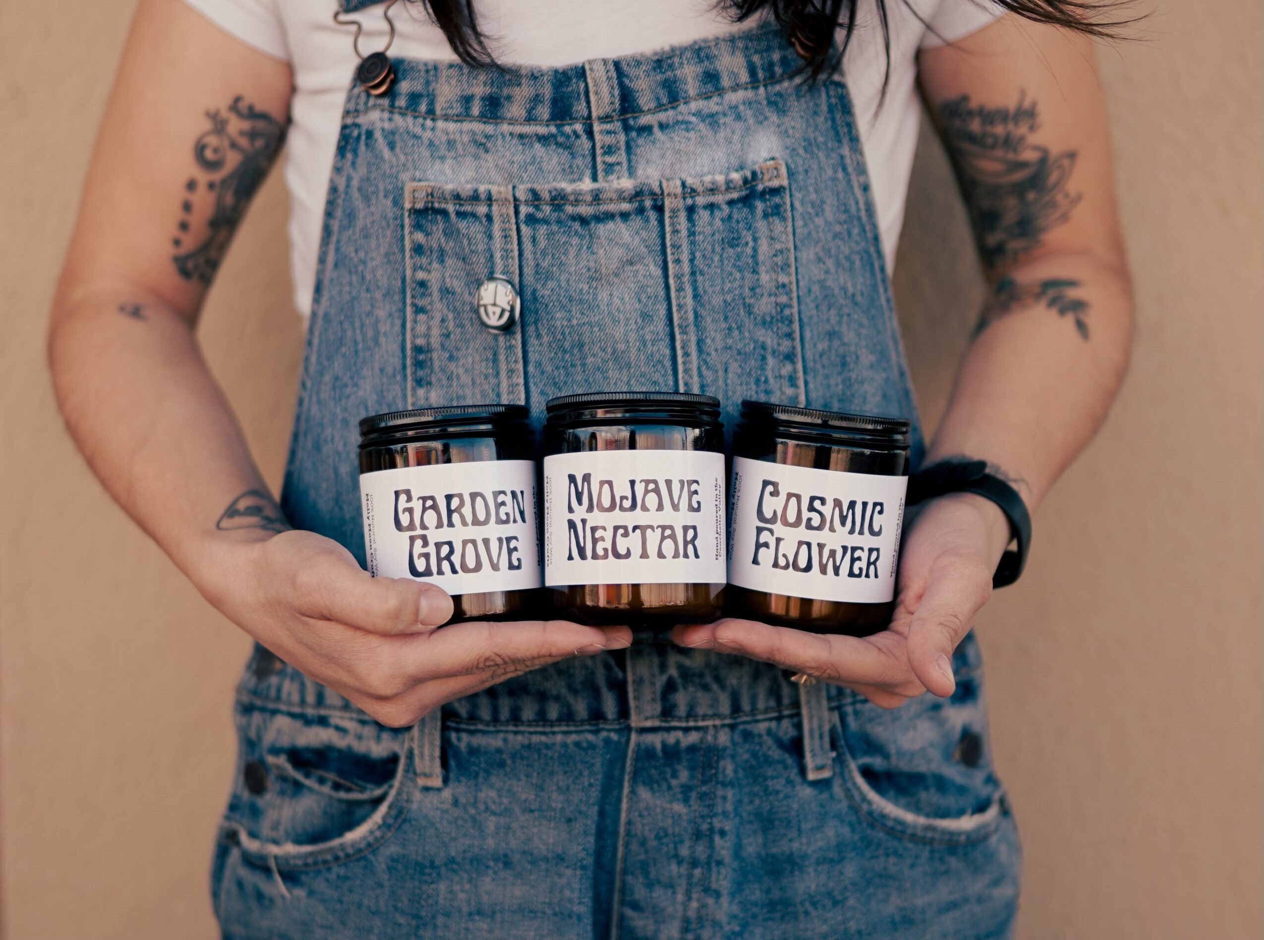 Person holding three jars labeled Garden Grove, Mojave Nectar, and Cosmic Flower, wearing denim overalls and a white shirt.