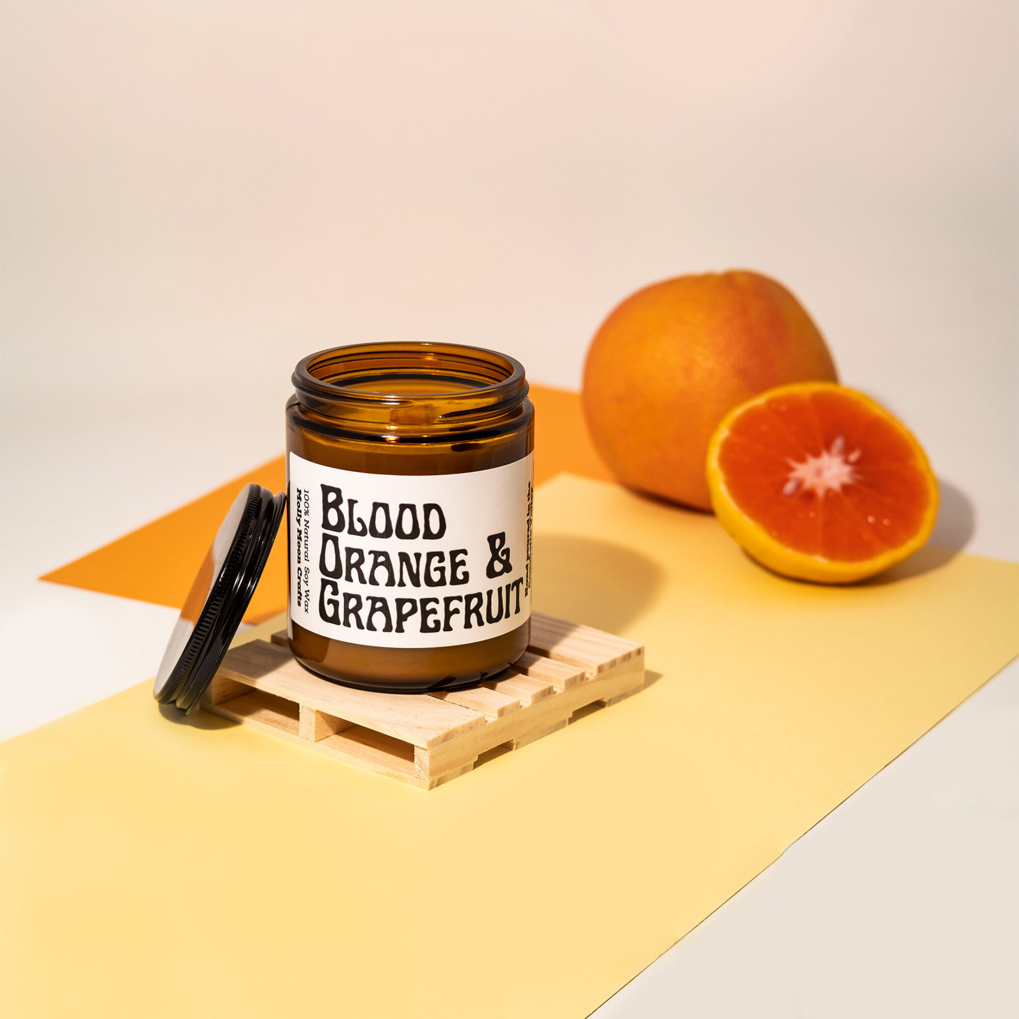 Open jar labeled 'Blood Orange & Grapefruit' with a whole and a halved blood orange beside it, on a yellow surface.