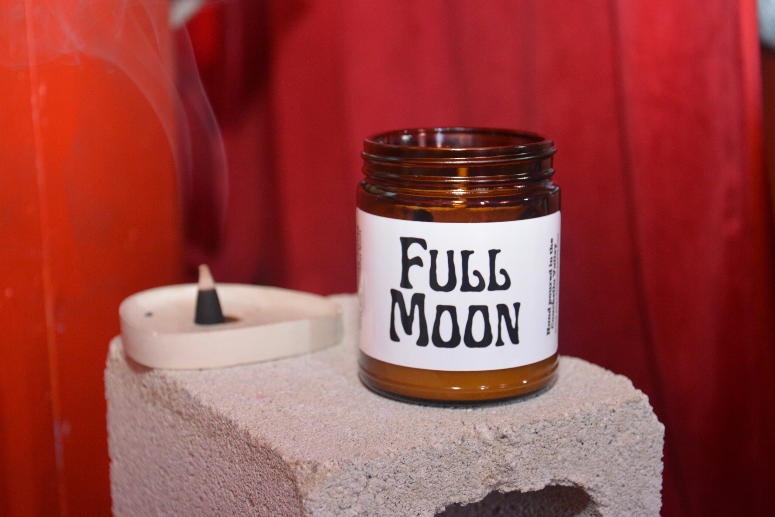 Open jar labeled 'Full Moon' on concrete block with candle and red background, no people visible.