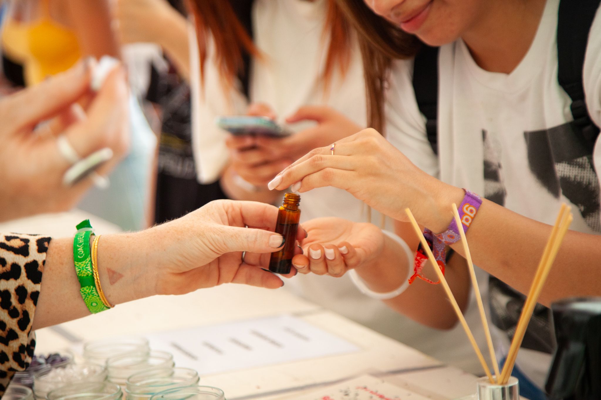 People exchanging small bottles at a table with papers and skewers, some using smartphones, in a busy setting.