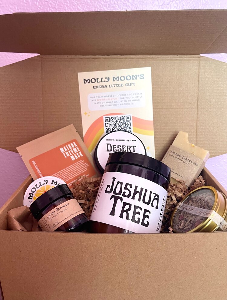 Open box containing jars, a card, a soap bar, and a small container, with a purple wall in background.