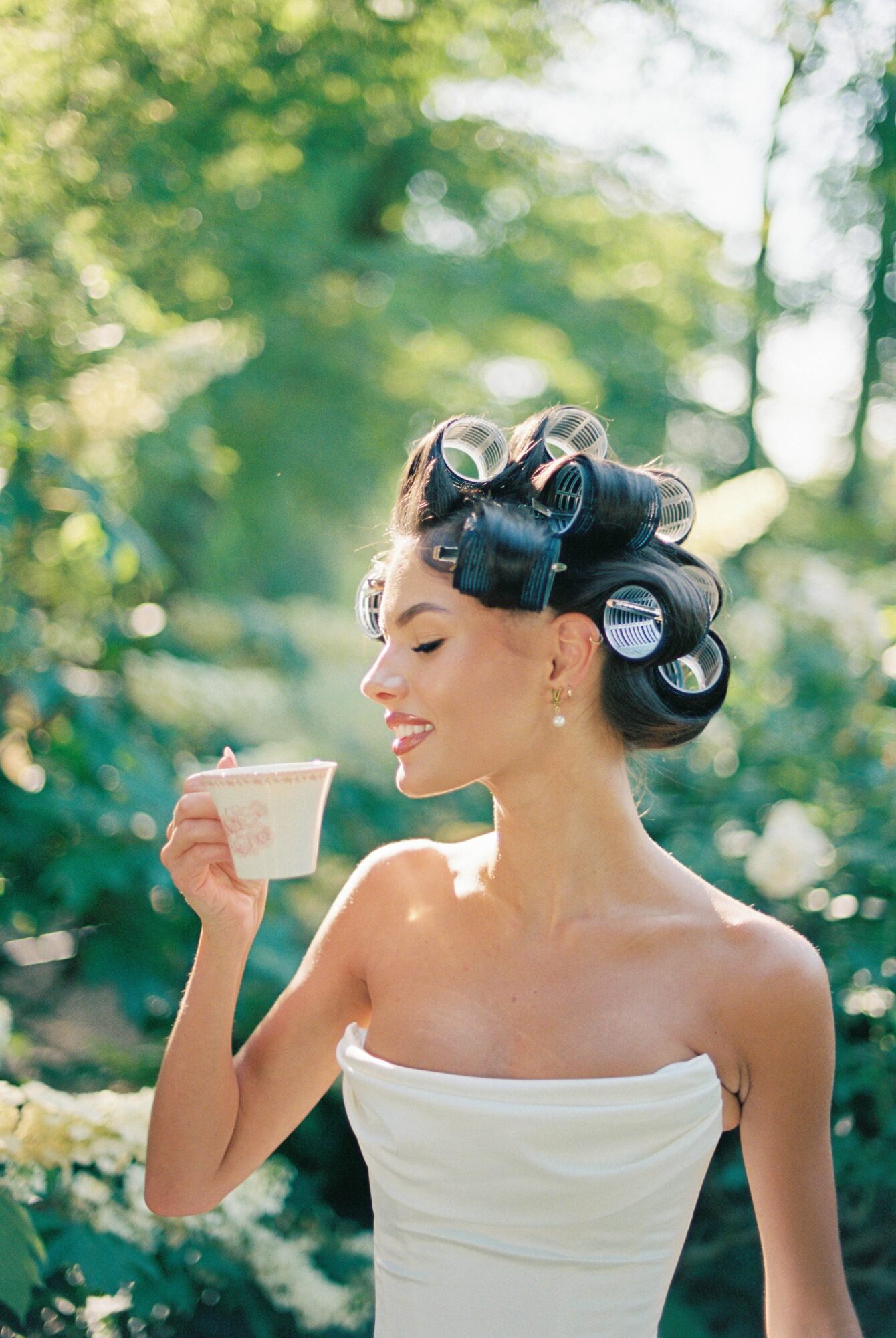 Woman with hair in rollers, holding a cup outdoors, smiling, wearing a strapless white top, trees in background.