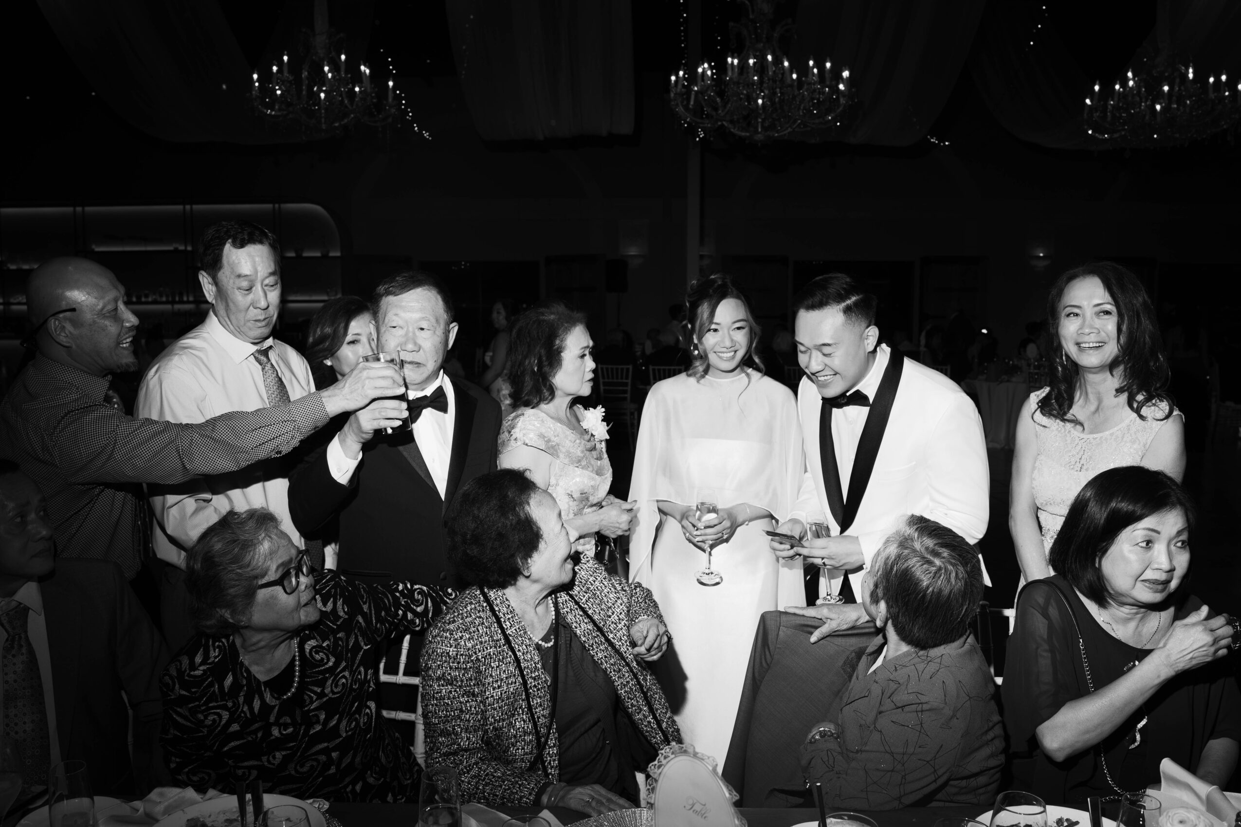 Group of people at a formal event, some holding glasses, smiling and engaging in conversation, in black and white.