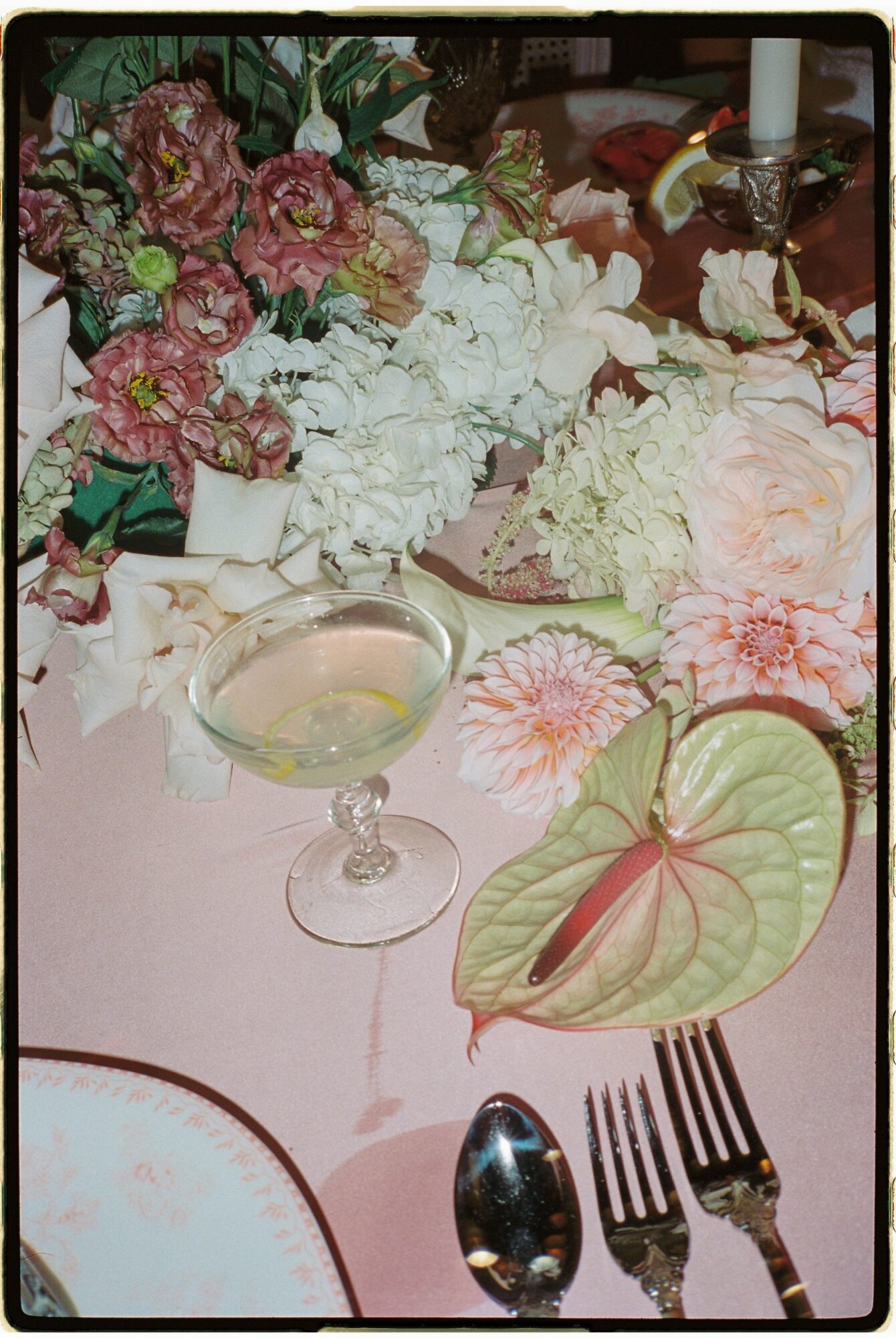 A floral centerpiece with pink and white flowers, a cocktail glass with a drink, and a plate with a green leaf and pink napkin, set on a pink tablecloth.