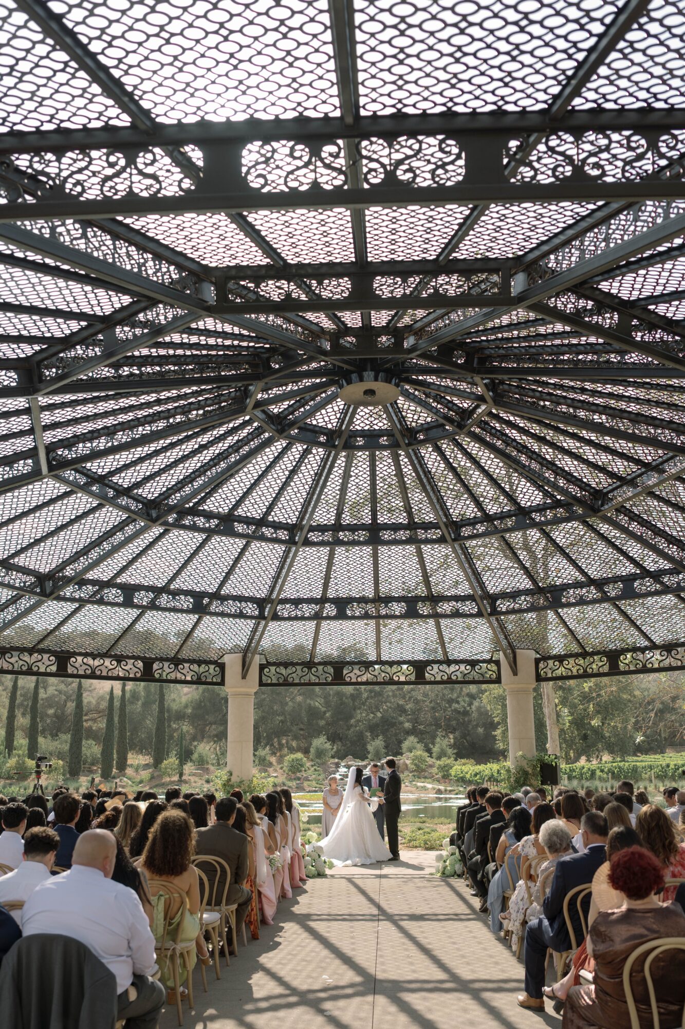 Wedding ceremony under a large ornate metal and glass dome with guests seated outdoors, couple in the center, bright daylight.