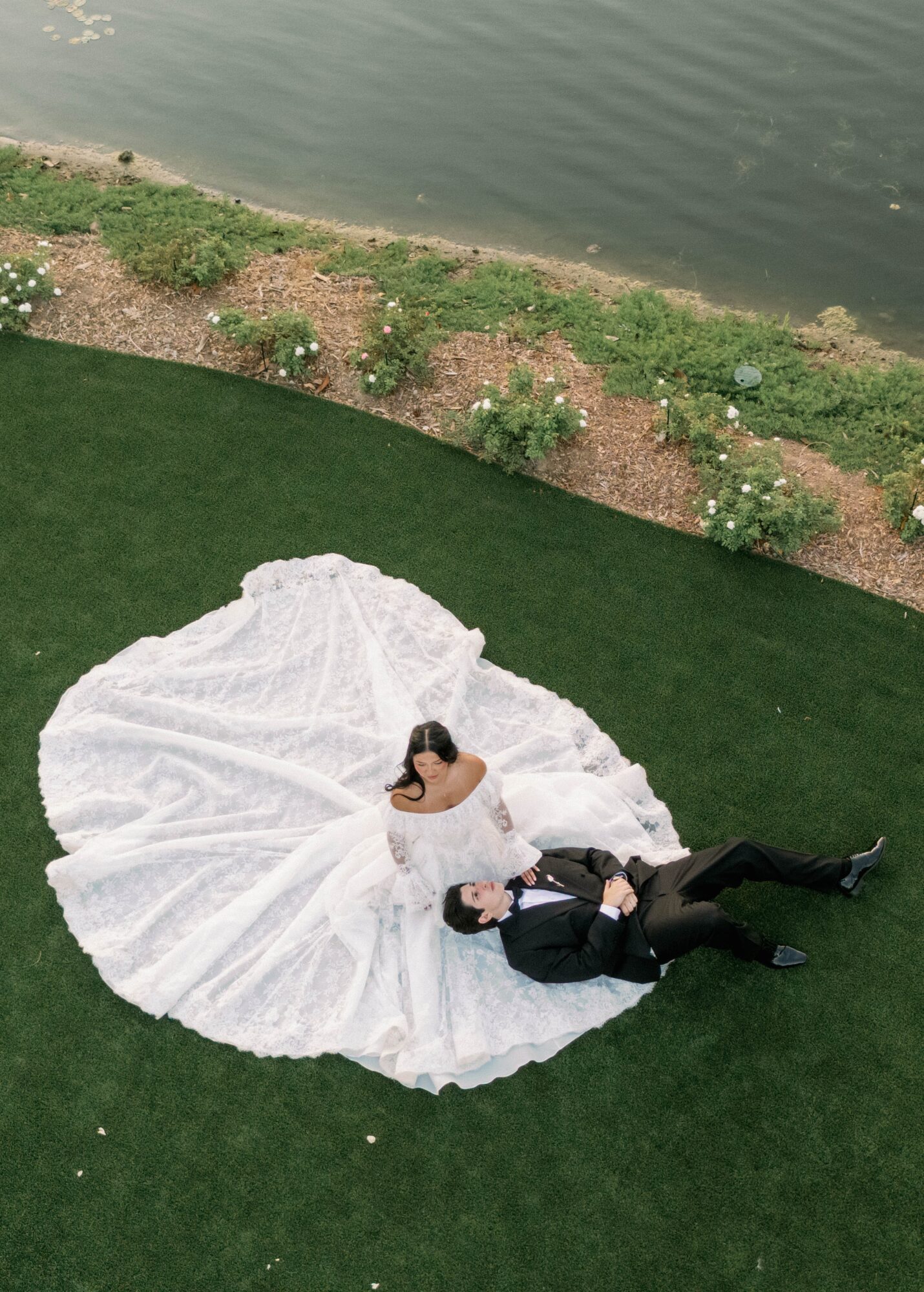 A bride in a white wedding dress and a groom in a black suit lie on grass near water, with flowers nearby.