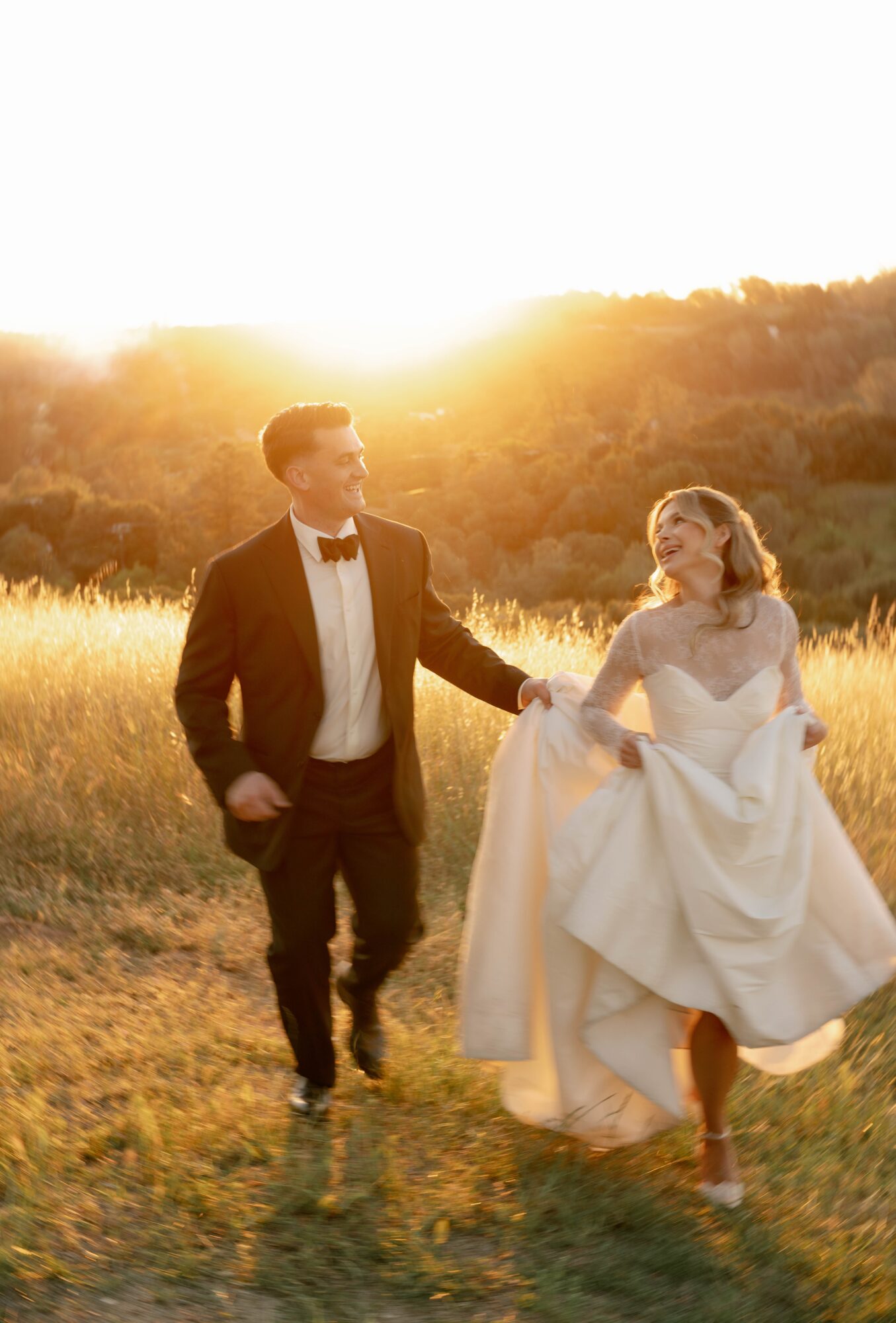 A couple walking in a field during sunset, the man in a tuxedo and the woman in a wedding dress, holding hands.