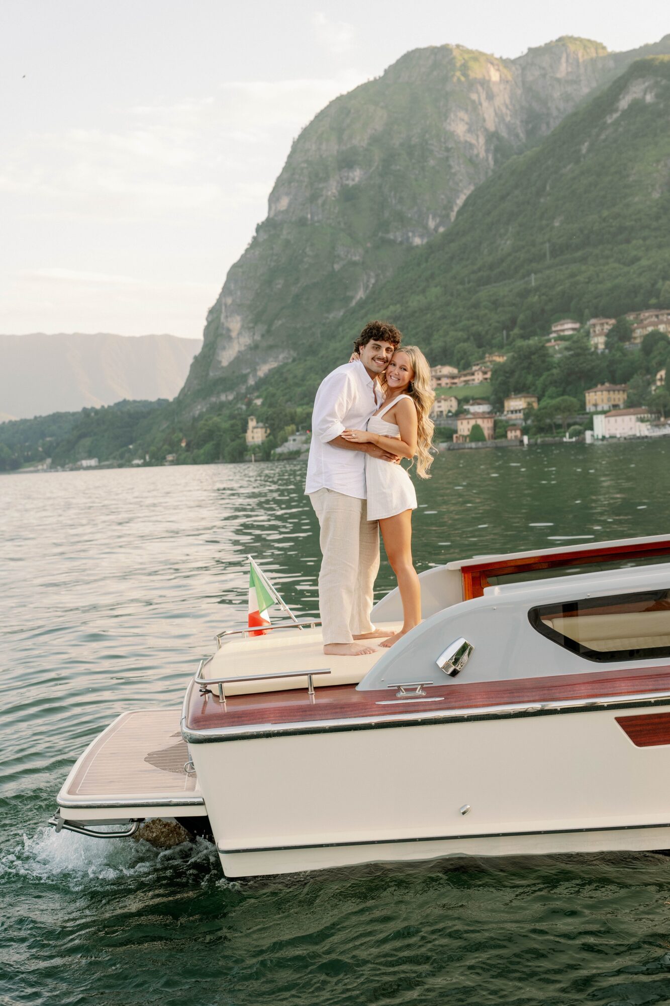 A couple hugging on a boat with a mountain and lake in the background.