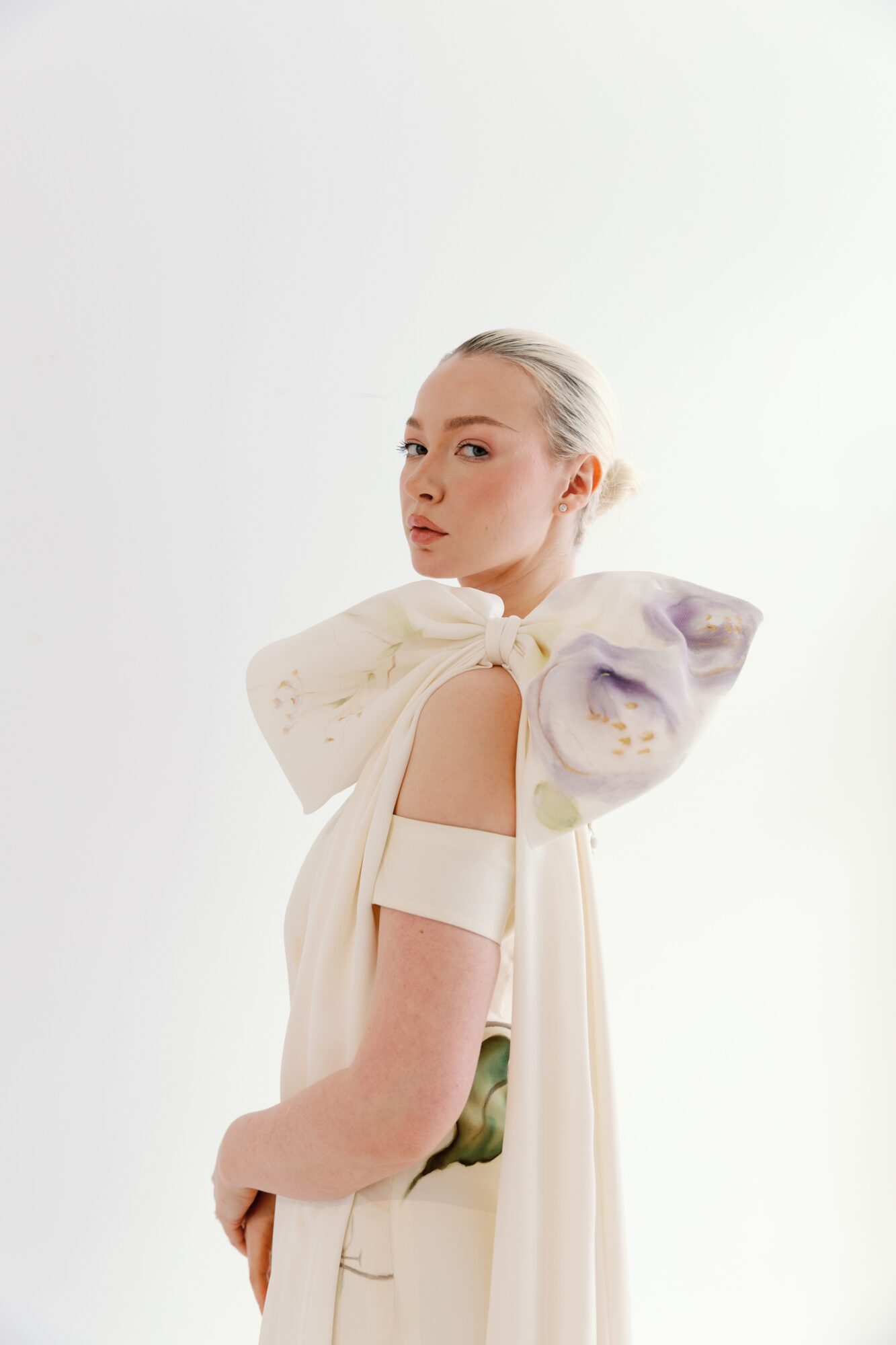 Young woman with platinum blonde hair in a high bun, wearing a white dress with large, colorful fabric bows on shoulders, standing against a plain white background.
