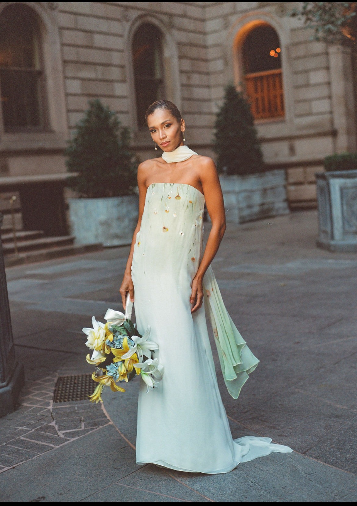 Young woman in a strapless white dress holding a bouquet of flowers, standing outdoors in front of a building with arched windows.