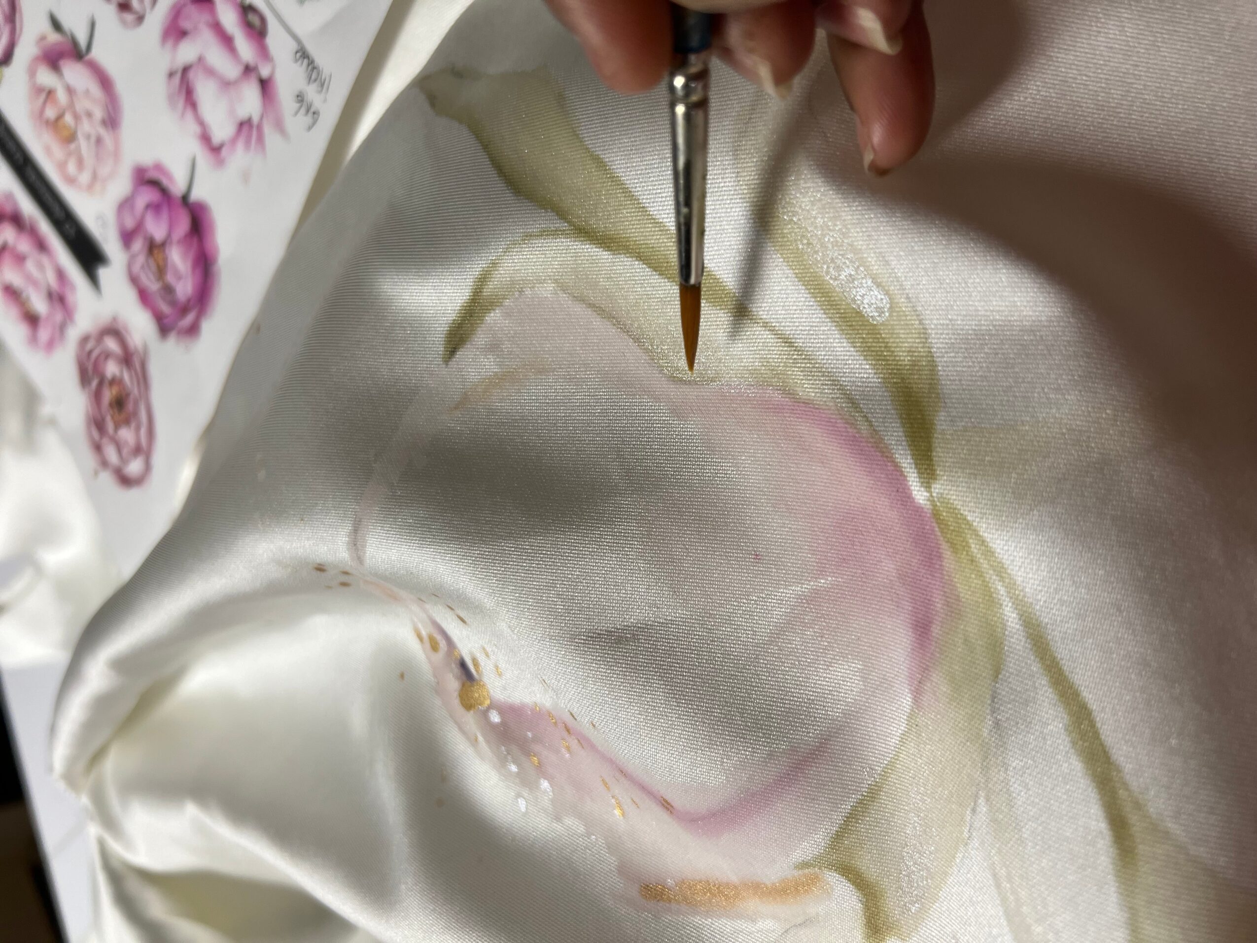 Person painting floral design on fabric with a fine brush, pink and green colors visible.