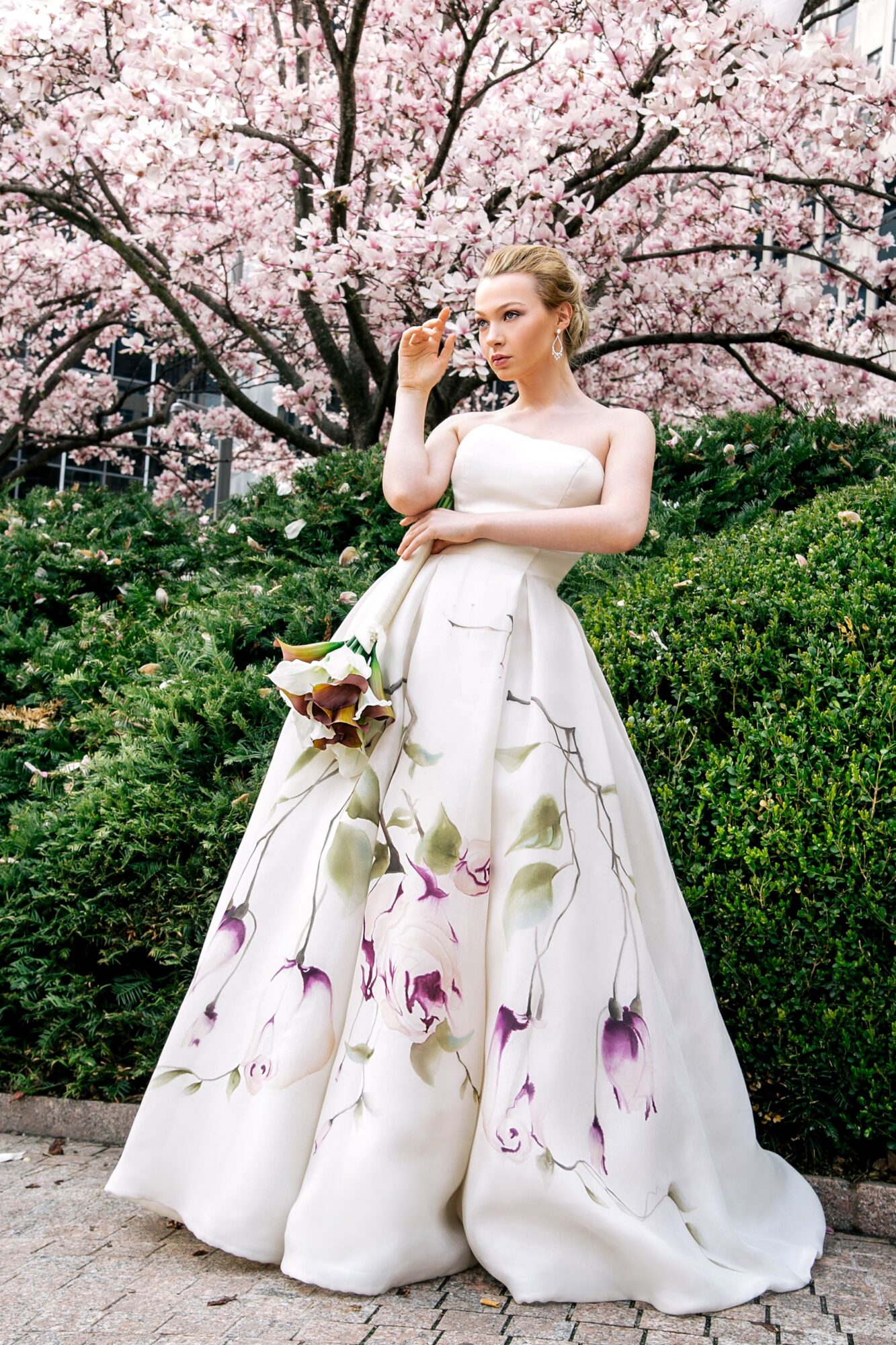Woman in a white floral gown standing outdoors with pink cherry blossoms in background.