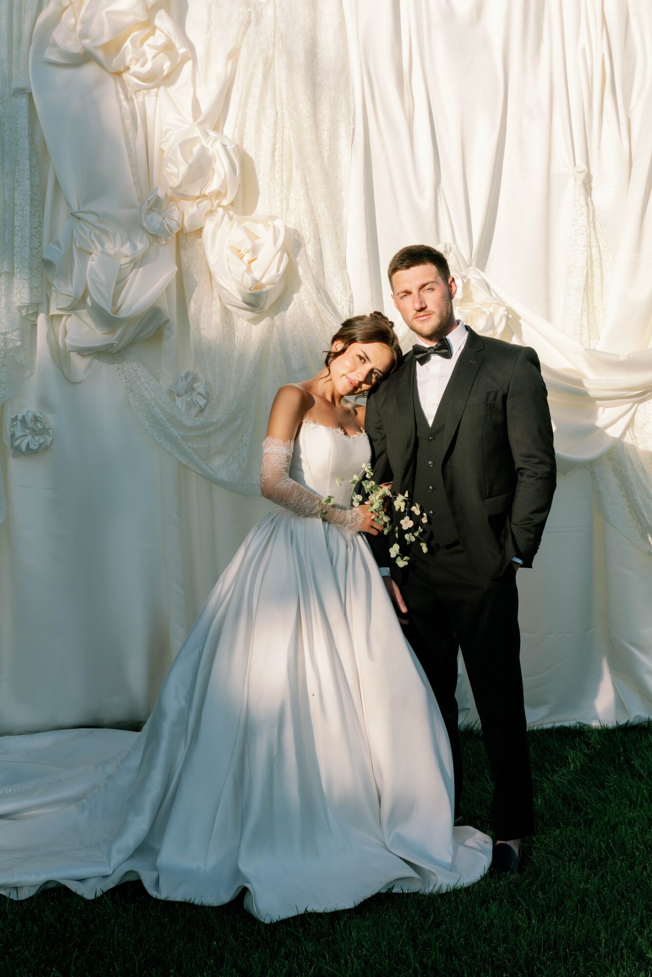 Bride and groom standing together in front of a white curtain backdrop, the bride leaning her head on the groom's shoulder, holding a bouquet.