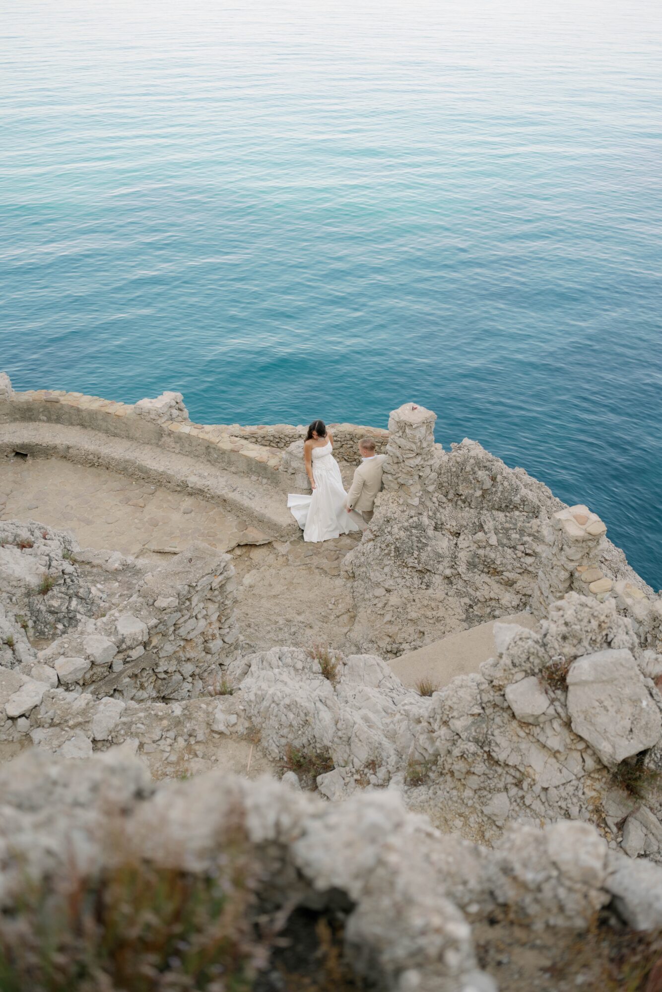 Person in white dress standing on rocky cliff overlooking calm blue sea, landscape with rocks and water, daytime.