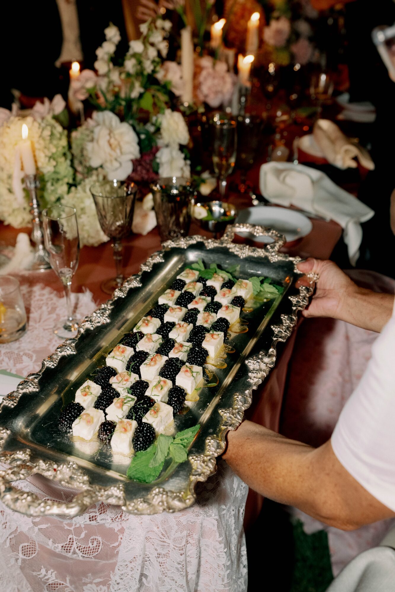 Person holding a silver tray with sushi rolls at a decorated table with flowers and candles, in a formal setting.
