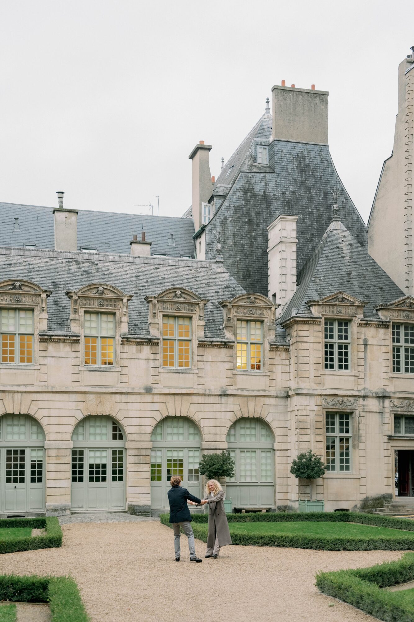 Two people stand and talk in front of a historic building with arched windows and a slate roof.