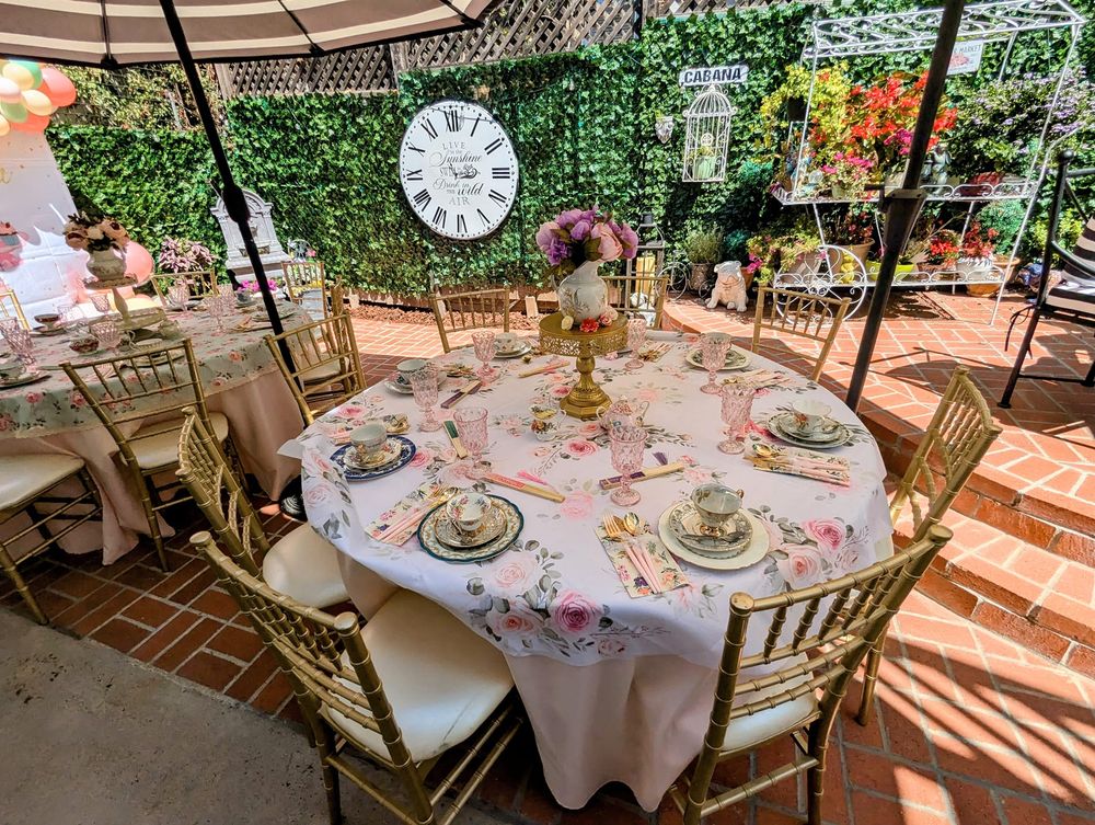 Round table with floral tablecloth, set with teacups, plates, and a centerpiece of pink flowers, outdoors with greenery and clock in background.