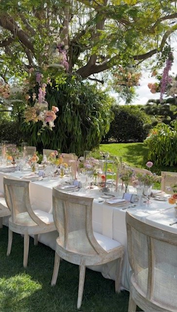 Outdoor garden setting with a long table, white chairs, and lush greenery, including flowering trees and bushes.