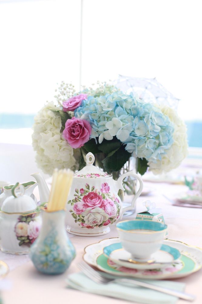 Floral arrangement with pink roses and blue hydrangeas on a table with teacups and decorative items.
