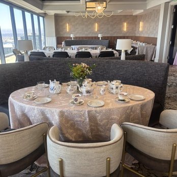 Round table with a floral centerpiece, set with teacups, saucers, and plates, in a bright, modern dining room with large windows.