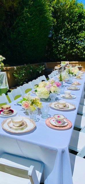 Long table set outdoors with white tablecloth, plates, and floral centerpieces, surrounded by white chairs, greenery in background.