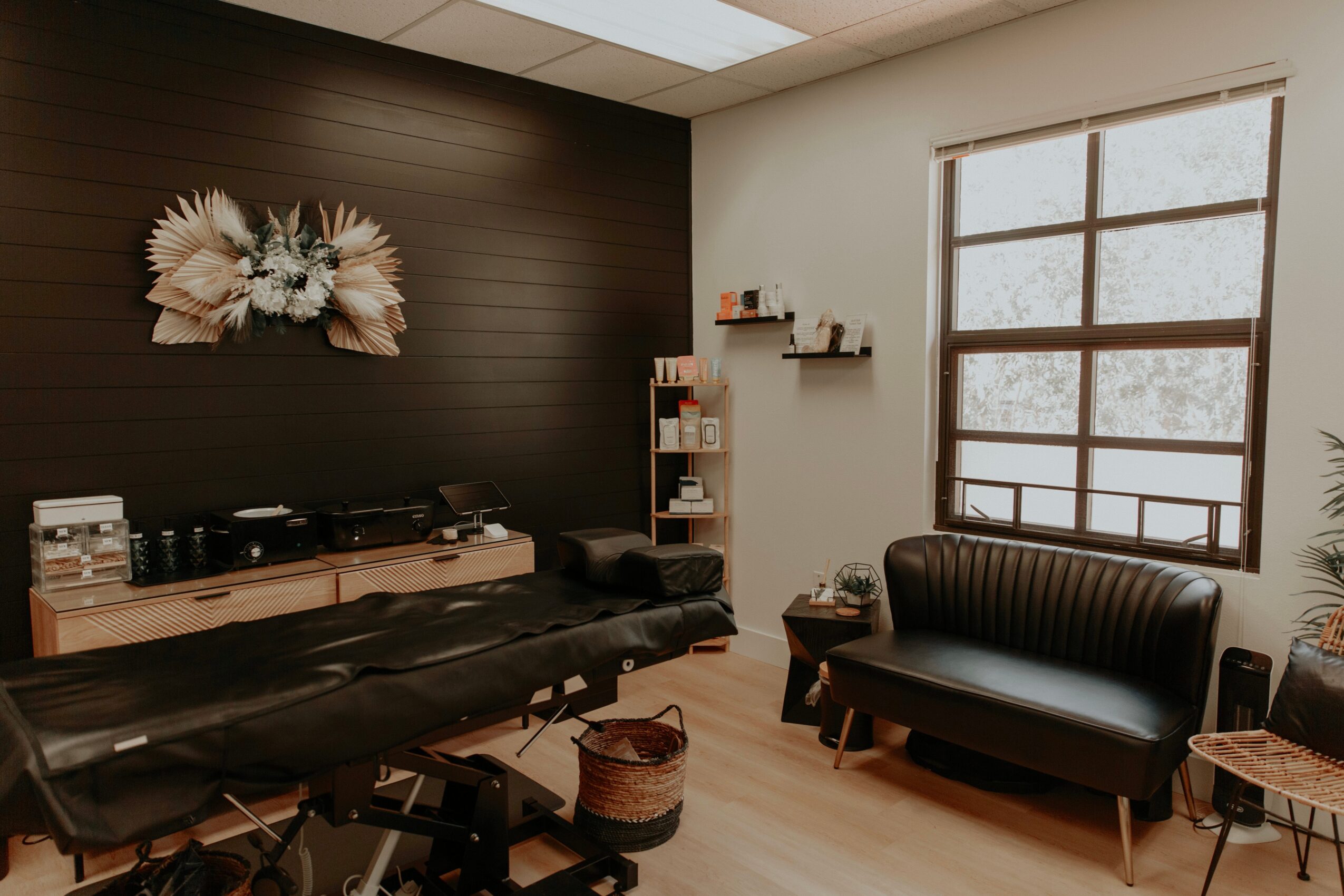 Room with dark wall, floral wall art, black sofa, window, and shelves, with light wood flooring and natural light.
