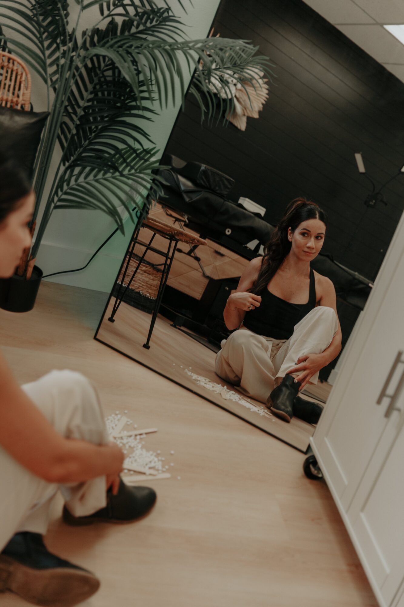 A woman sits on the floor in front of a mirror, adjusting her hair, with a large plant nearby and a black wall behind.