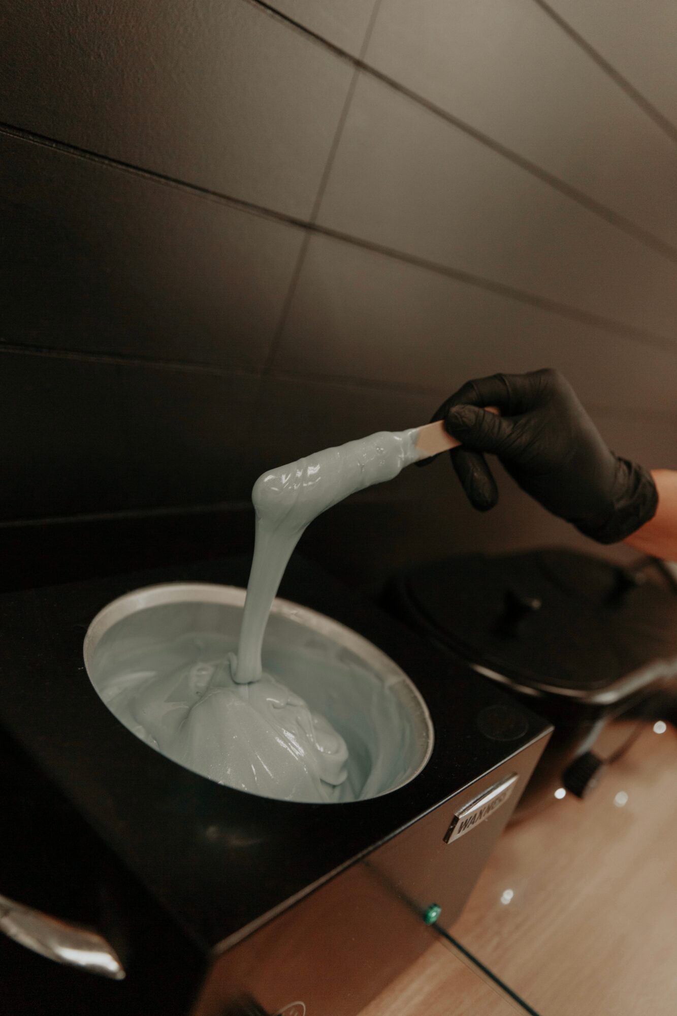 Person wearing a black glove pouring a white, viscous substance into a container, against a dark background.