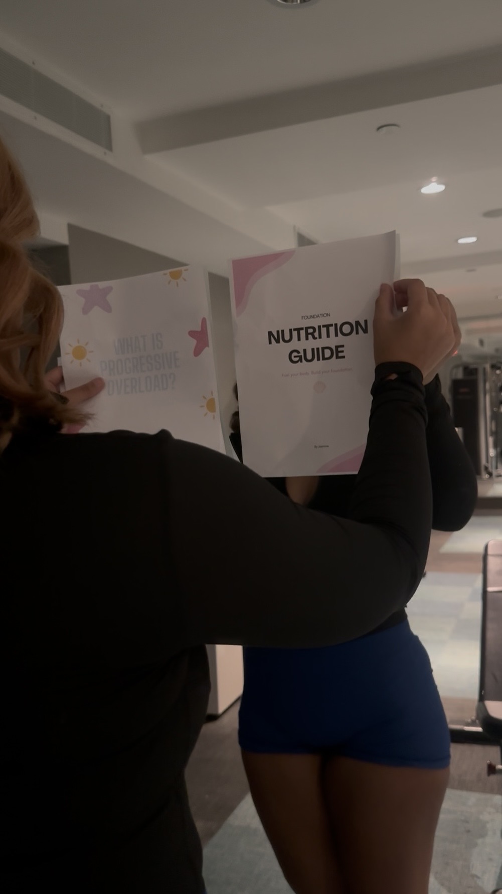 Person holding a nutrition guide in a public indoor space, with a person in blue shorts nearby.