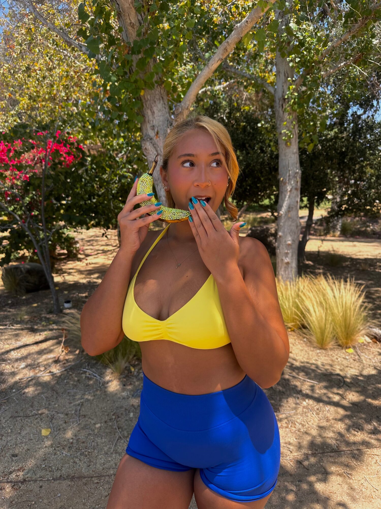 Woman in yellow top and blue shorts holding a phone and a snack outdoors with trees and plants behind her.