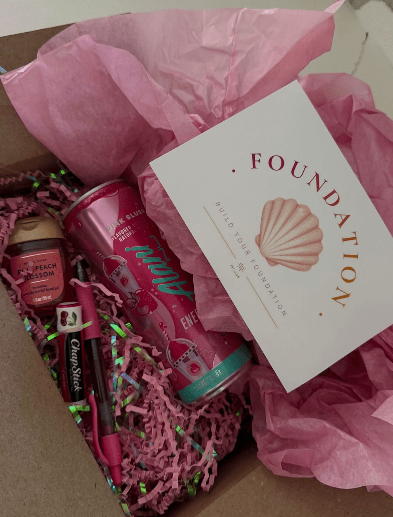 Gift box with pink tissue paper, a card with a seashell, and various pink items inside.