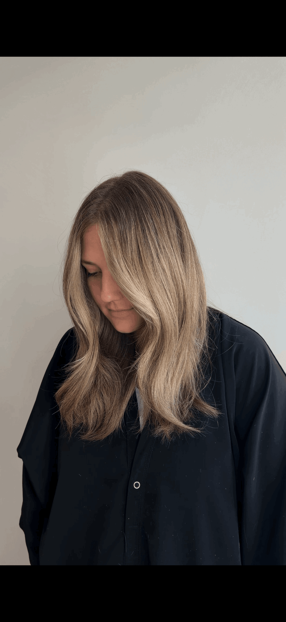 Person with long, wavy blonde hair looking downward, wearing a black shirt, against a plain light background.