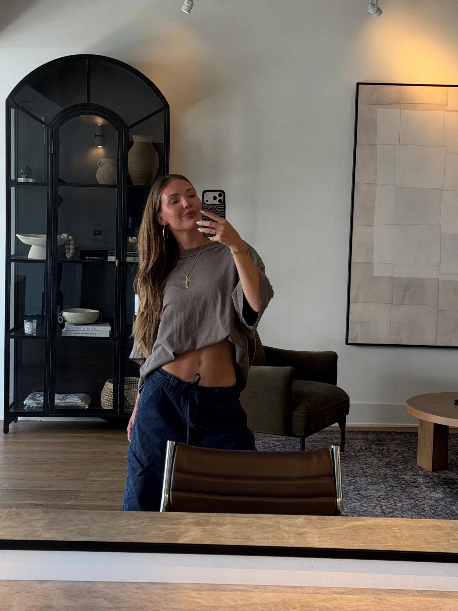 Young woman taking a mirror selfie in a modern living room, wearing a gray top and dark pants.