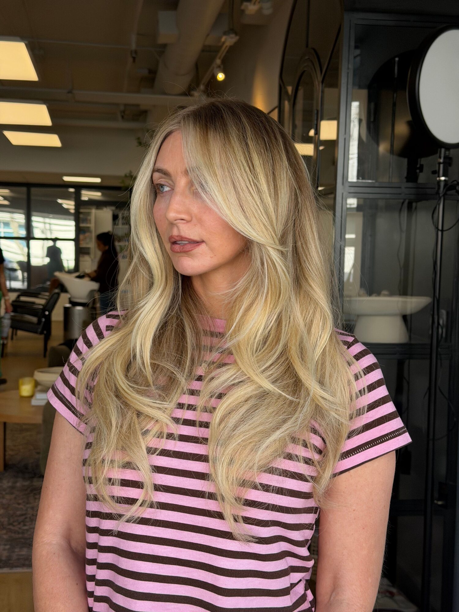 Woman with long blonde hair wearing a pink and white striped shirt, looking down, in an indoor setting.