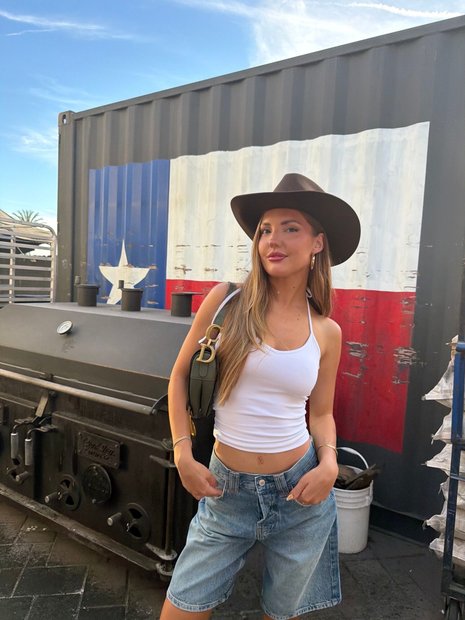 Woman wearing a black hat, white tank top, and jeans, standing outdoors near a colorful wall.