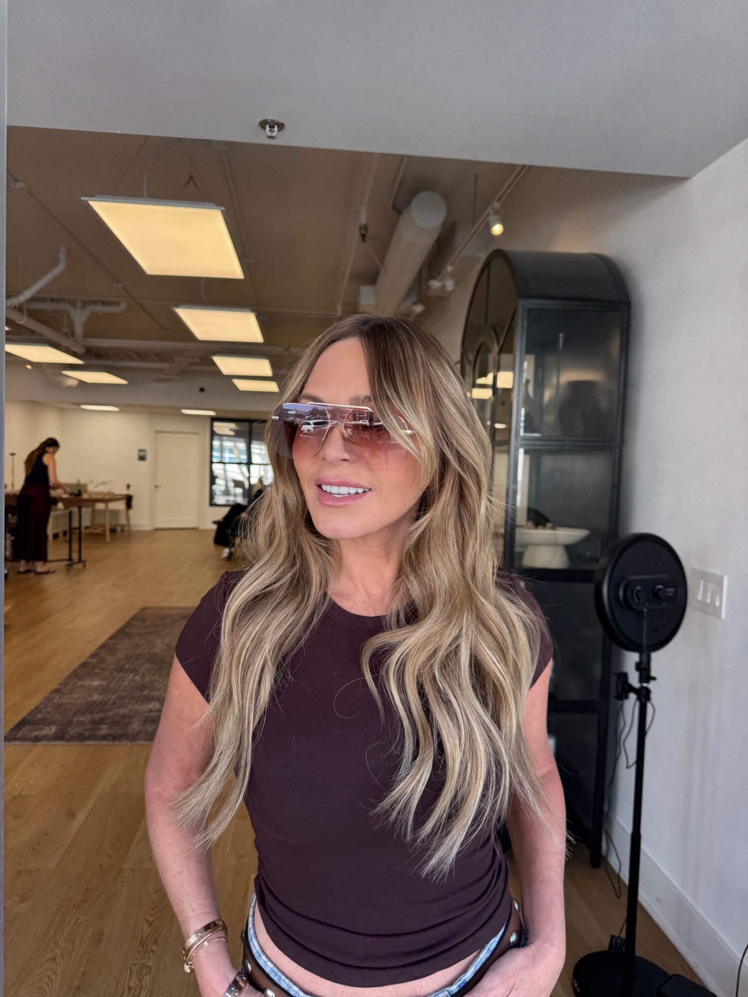 Woman with long wavy blonde hair wearing glasses and a dark top, smiling in a room with wooden floors and a kitchen in the background.