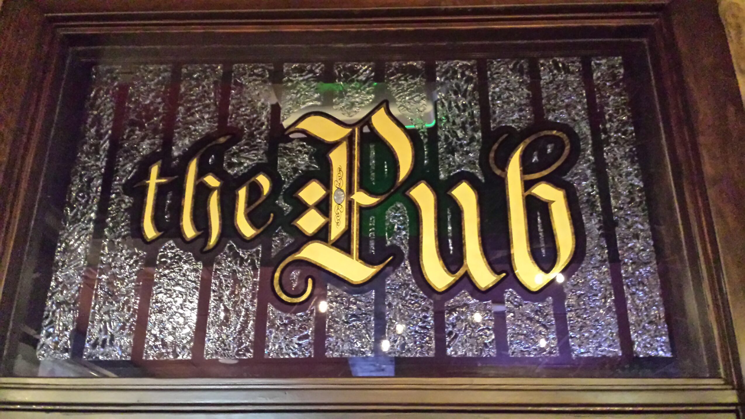 Sign with the words 'the Pub' in black and gold Gothic font, behind a metal gate with a background of trees.