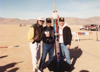 Three people standing outdoors near a tall object in a desert landscape with vehicles in the background.
