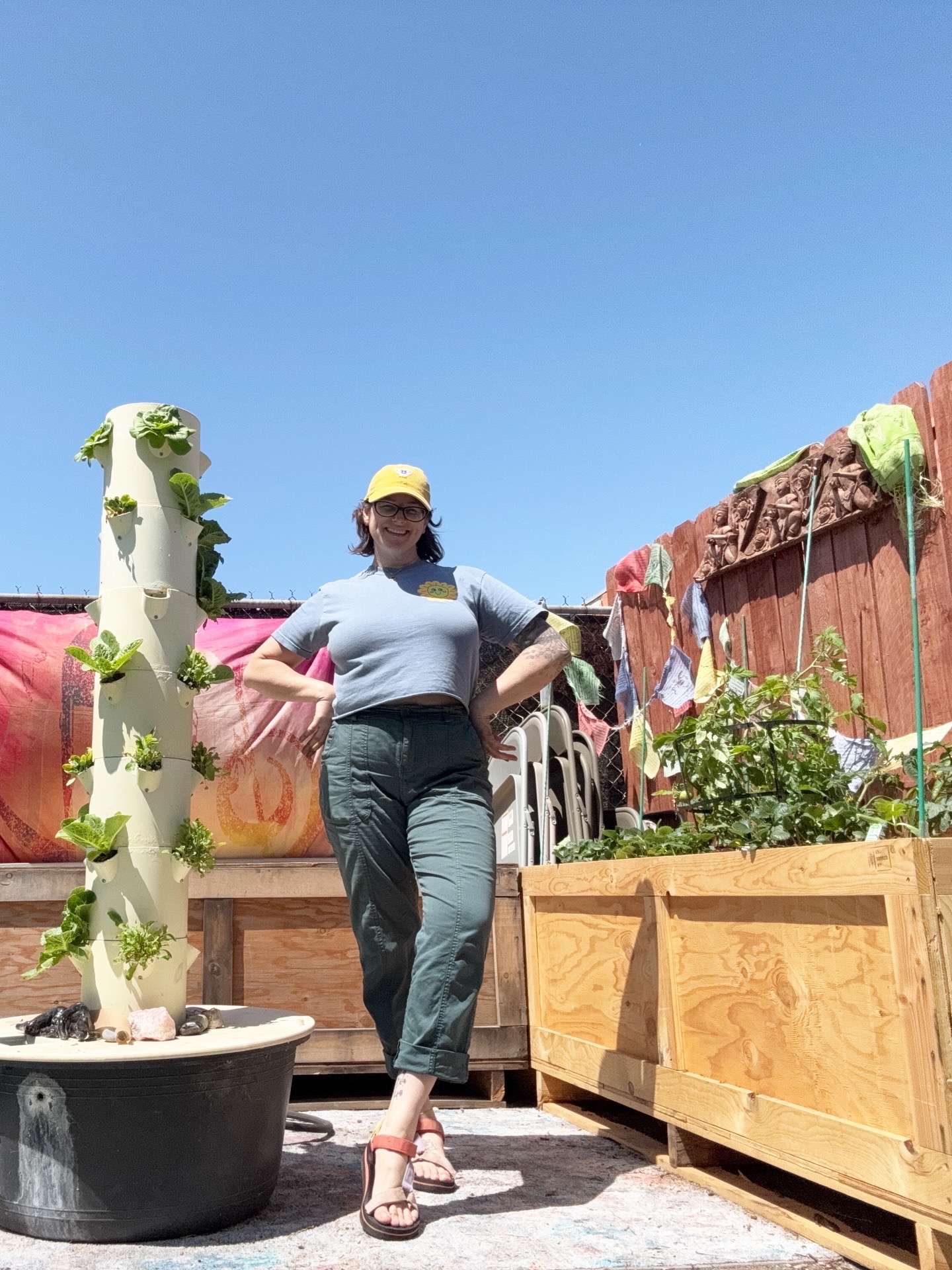Woman standing outdoors with arms on planters, wearing a yellow cap, gray shirt, and dark pants, against a clear blue sky.