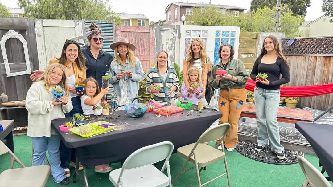 Group of women and children gathered around a table outdoors, celebrating a birthday with gifts and decorations.