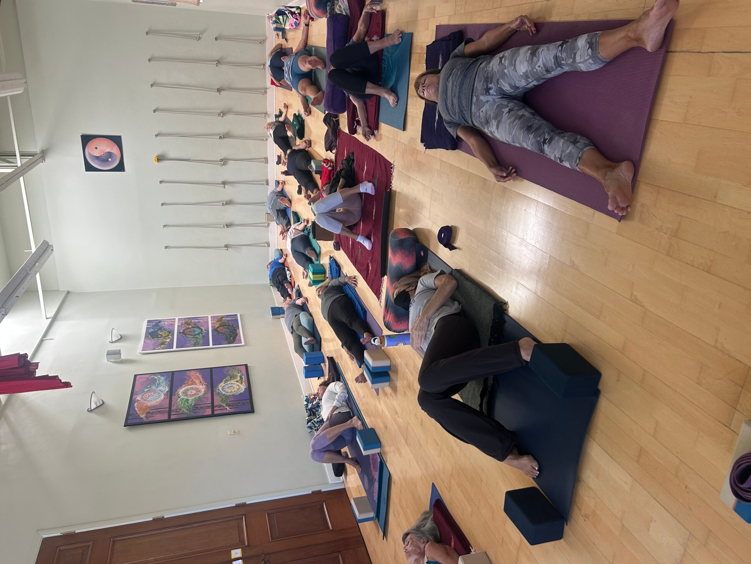 People sitting on yoga mats in a room, participating in a group activity or class, with artwork on the wall.