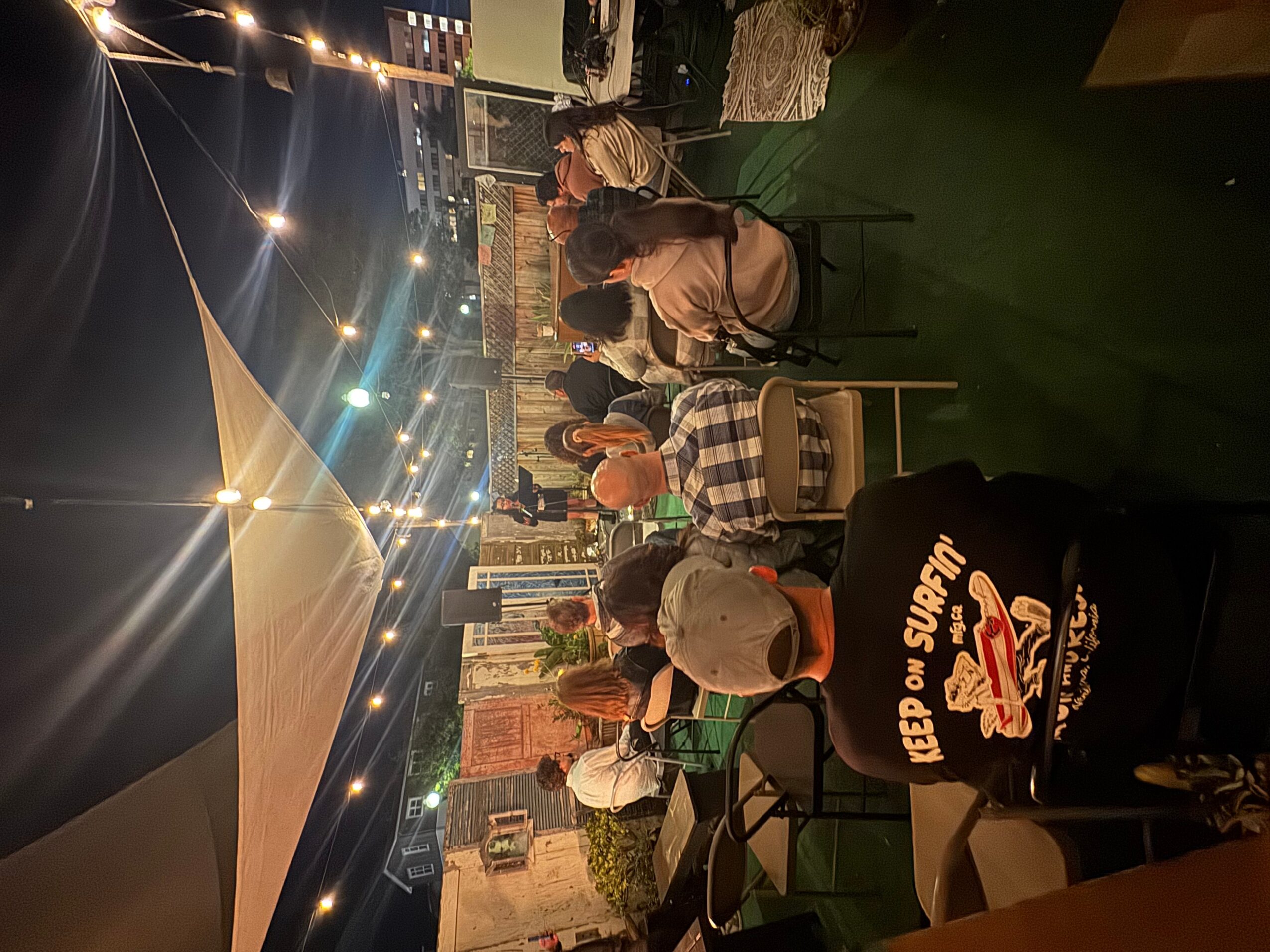 People seated at tables outdoors under a large canopy at night, with string lights overhead and a stage in the background.