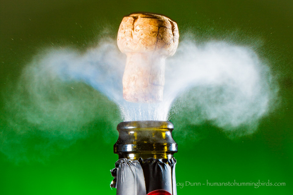 A glass bottle with a cork releasing a cloud of vapor or smoke against a green background.