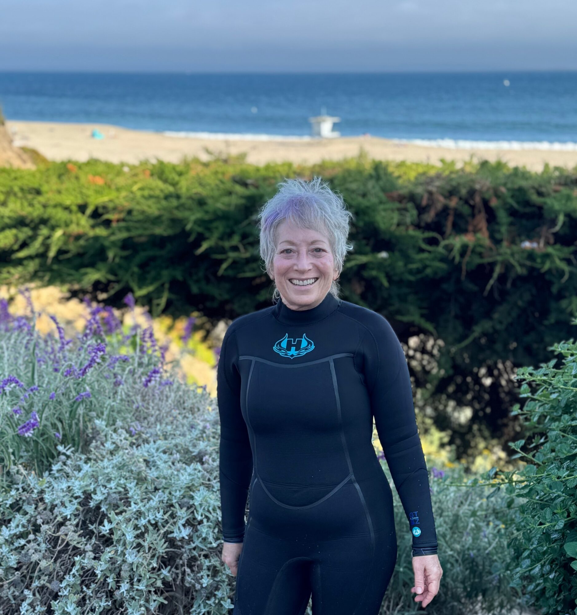 Smiling woman in a wetsuit standing outdoors with a beach, ocean, and greenery in the background.