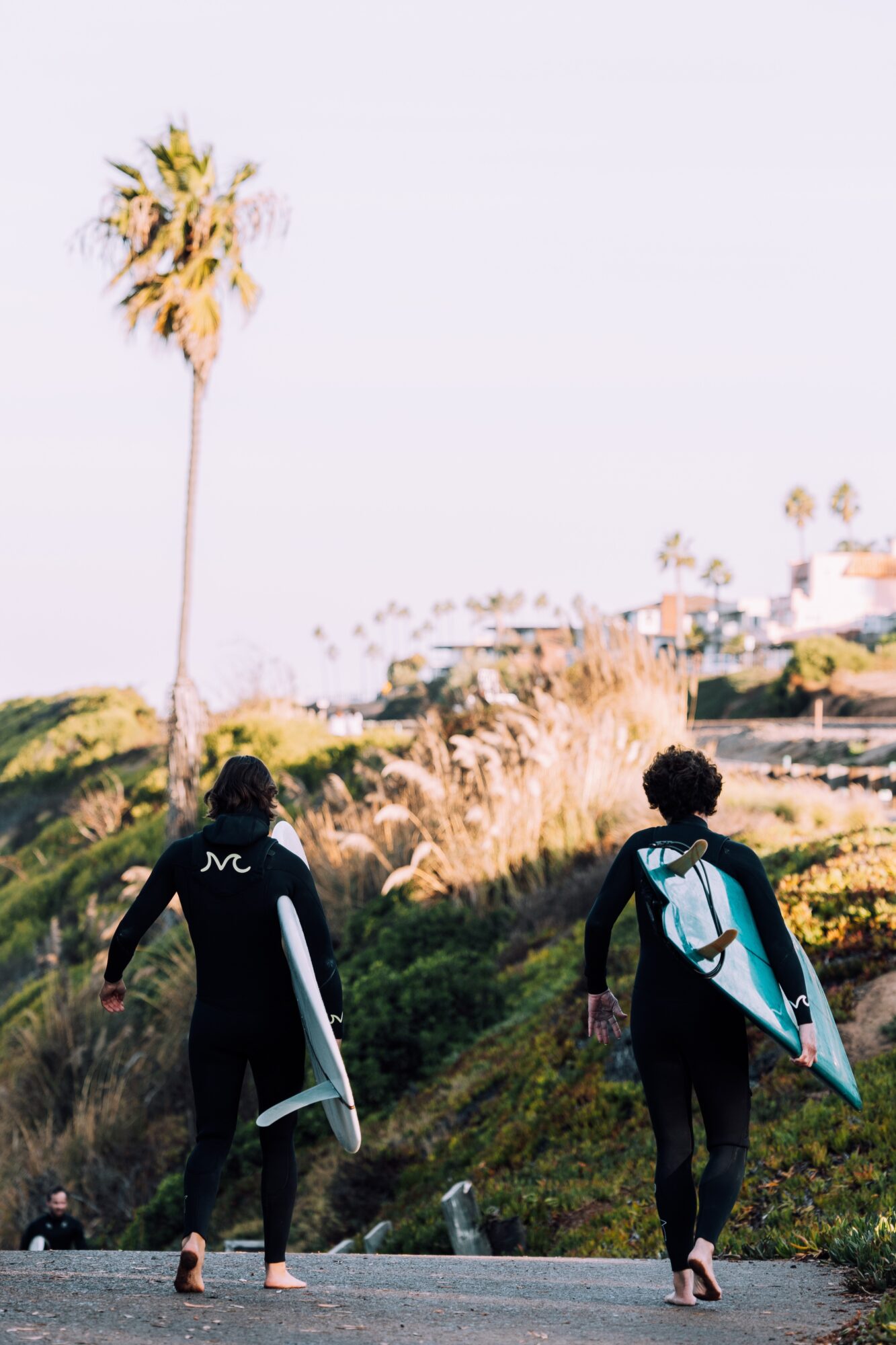 Two people in wetsuits walking with surfboards on a grassy path, palm trees in the background, clear sky.