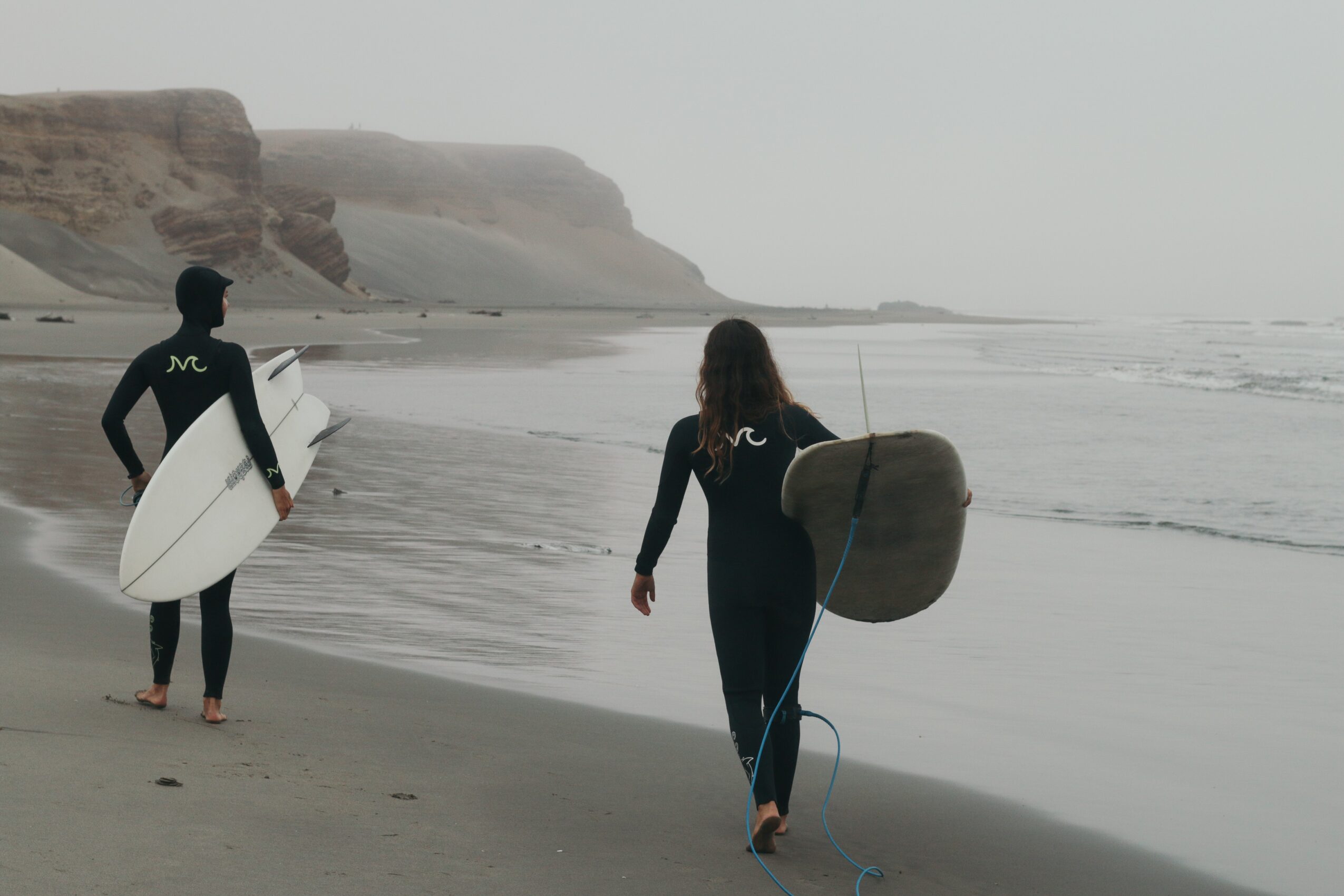 Two people with surfboards walk along a beach with cliffs in the background, overcast sky, and calm ocean waves.