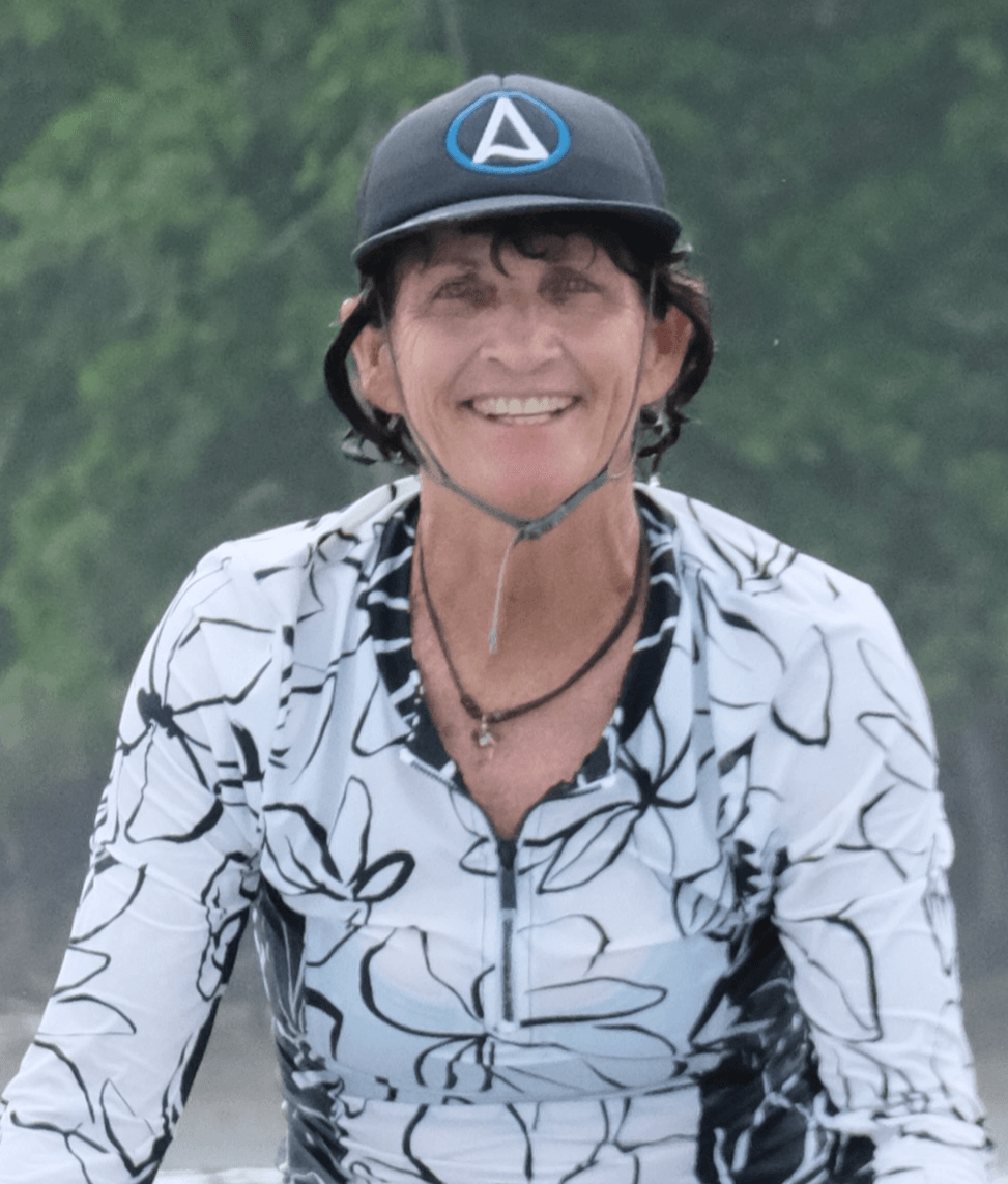 Smiling woman wearing a helmet and patterned shirt outdoors, with trees in the background.