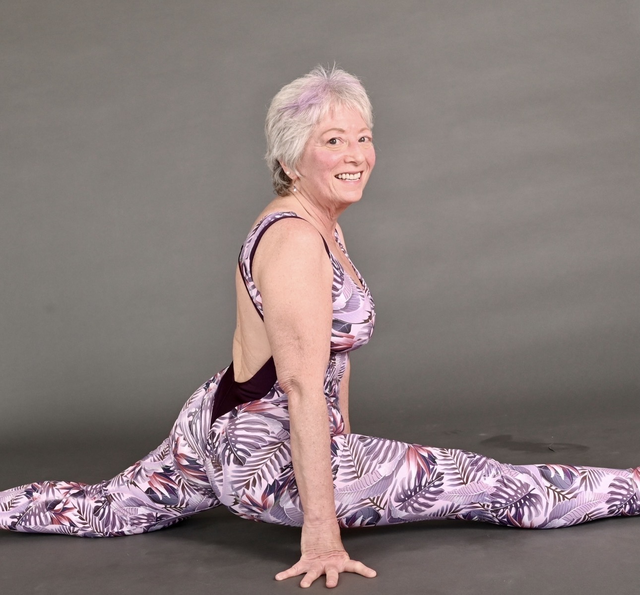 Older woman performing a split stretch on a gray background, smiling, wearing a patterned workout outfit.
