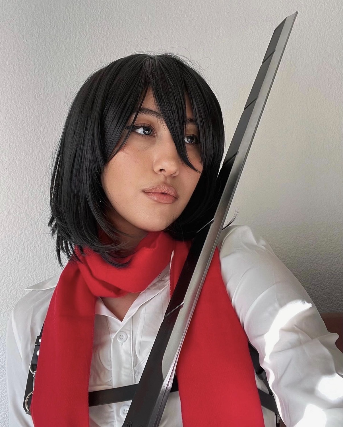 Woman with black hair, white shirt, red scarf, looking at a mirror, against a plain wall.