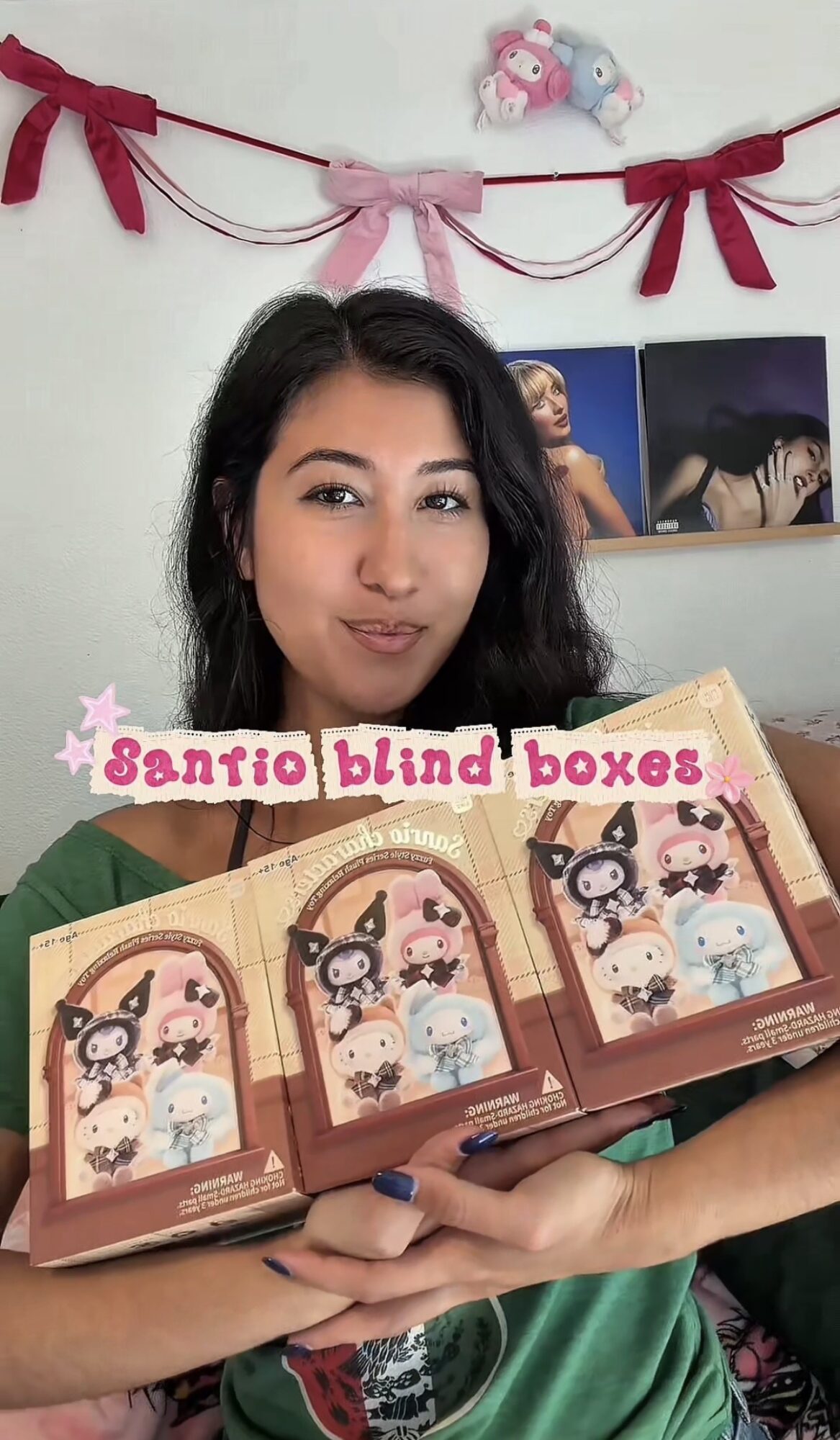 Young woman holding three boxed Santino blind boxes, smiling, with decorations and photos on wall behind her.